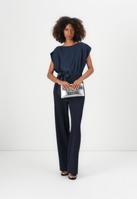 Top azzurro navy, a maniche corte con cintura a fiocco, abbinato a pantaloni lunghi navy. Il modello tiene una borsa a tracolla metallica argento. Texture liscia in tutto.