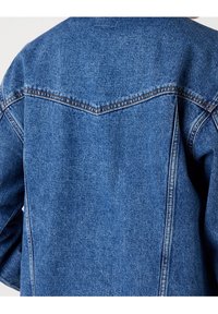 Džínová bunda v středně modré barvě, s viditelnými švy na hrdle a ramenech, s uvolněným střihem a měkkou texturou.