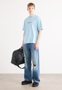 Lyseblå bomulls t-skjorte med rund halskant og logo på fronten, sammen med slitte, lyse blå denimjeaner. Svart veske og kraftige svarte sko.
