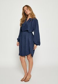 Temno modra obleka z ovratnikom na gumbe, dolgimi rokavi in pasom, ki se zaveže. Ima teksturirano tkanino in stranske zareze na robu.