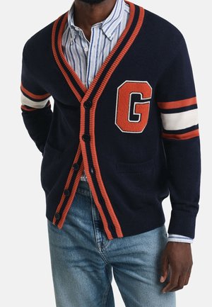 Mężczyzna w granatowym kardiganie z pomarańczowo-białymi paskami na rękawach i dużą pomarańczową literą "G" na piersi, w połączeniu z niebieskimi dżinsami.