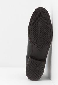 Chaussure en cuir noire avec une semelle en caoutchouc texturée présentant un motif en zigzag, un logo en relief et une étiquette de taille 37.