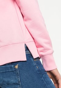 Vista lateral de una persona que lleva una camiseta de manga larga rosa con una abertura lateral y pantalones vaqueros azul denim que muestran costuras y un detalle de botón de metal.