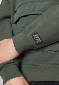 Sudadera verde con un bolsillo con cremallera, puños acanalados y una etiqueta en la manga. La tela parece tener textura y un diseño casual.