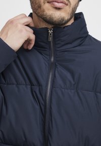Navy puffer jacket features a high collar, zip closure, and textured fabric with a subtle pattern. Soft and lightweight material.