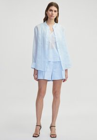 Helles blaues Leinenhemd mit Knopfleiste und Mandarin-Kragen, drüber getragen über einem Oberteil mit Spitze und kombiniert mit passenden Shorts.