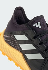 Czarny sportowy but z teksturowaną cholewką, pomarańczową podeszwą środkową, białym logo w trzy paski oraz różowym przeszyciem z boku.