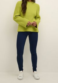 Jasnozielony sweter z prążkowanej tkaniny o szerokich rękawach, zestawiony z ciemnoniebieskimi skinny jeansami i białymi trampkami. Widoczna lekka struktura materiału.