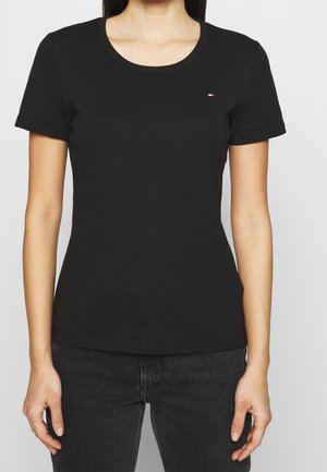 Frau trägt ein enganliegendes schwarzes T-Shirt mit kurzen Ärmeln, einem kleinen rechteckigen Logo auf der linken Brust und dunklen Jeans.