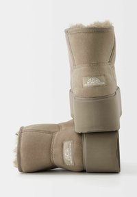 Béžové kotníkové boty vyrobené z semiše s chlupatým vnitřkem. Mají plochou, silnou podrážku a logo na boku. Viditelné texturované prošívání.