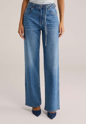 Femme portant un jean bleu taille haute à jambes larges avec une fine ceinture en denim nouée à la taille et des chaussures pointues bleu marine.