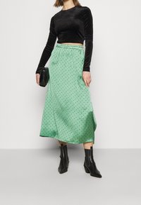 Kvinna i svart långärmad crop top, grön midiklänning med prickar, svarta ankelstövlar och håller en svart kuverttväska.