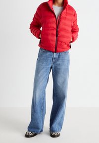 Rød pufferjakke med zip foran, rund krave og quiltet tekstur. Sammen med bredbenede lyseblå jeans og stribede slip-on sko.