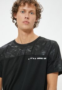 Sort atletisk t-shirt med en tekstureret, camouflage-mønstrede øverste del og teksten "LEGEND INSIDE YOU." i hvid.
