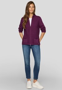 Lila Zip-Cardigan mit geripptem Muster, ausgestattet mit zwei Fronttaschen. Kombiniert mit blauen Jeans und weißen Turnschuhen.