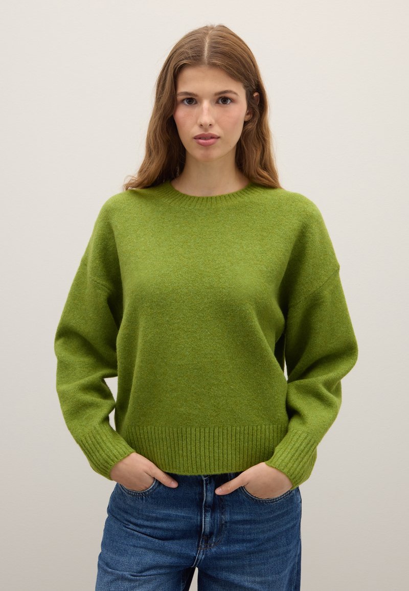 Suéter verde de punto grueso con cuello redondo, hombros caídos y puños y dobladillo acanalados. Combinado con vaqueros azules de mezclilla.