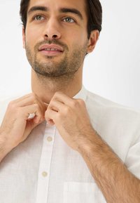 Camicia bianca di lino con colletto a collo alto. La camicia presenta una chiusura a bottoni e una tasca sul lato sinistro. Il modello sta allacciando il colletto.