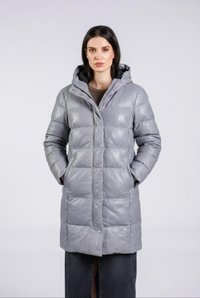 Kvinde iført en lang, lys grå polstret vinterfrakke med hætte, hænderne i lommerne, stående mod en ensartet lys baggrund.