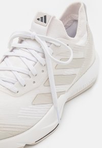 Zapatilla deportiva blanca con tela texturizada, acentos de plástico y un collar acolchado. Incluye cordones blancos planos y un logo en relieve en la lengüeta.