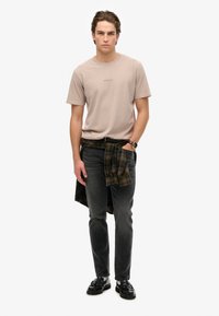 Beige bomulds T-shirt med rund hals, parret med mørke grå jeans og et grønt ternet flannel skjorte bundet om livet. Sorte sko fuldender looket.