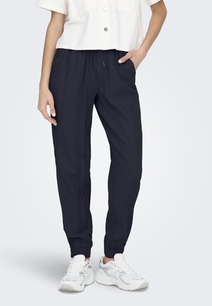 Persona que lleva pantalones jogger negros con puños elásticos, zapatillas blancas y una camisa blanca de manga corta con botones, mano en el bolsillo.