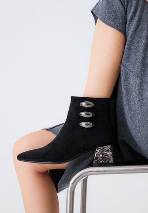 Botín negro de ante hasta el tobillo con tres botones metálicos y un exclusivo tacón de bloque texturizado. El diseño es puntiagudo en la puntera.