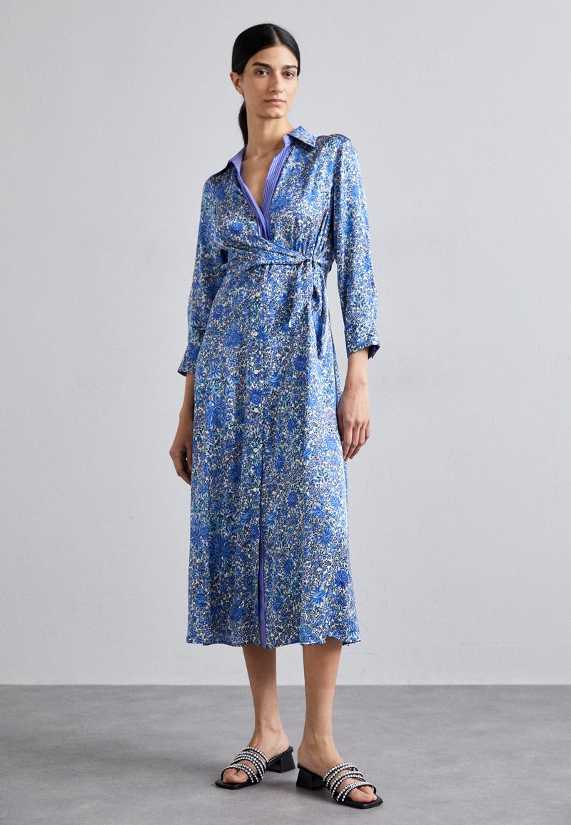 Blaues Blumen-Wickelkleid mit langen Ärmeln und Kragen, ausgestattet mit einem taillierten Gürtel und kontrastierenden lila Akzenten, kombiniert mit schwarzen Sandalen.