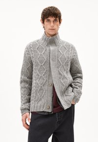 Graue Strickjacke mit hochgeschlossenem Kragen, die über Diamantmuster, eine Knopfleiste und gerippte Bündchen verfügt. Über einem dunklen Oberteil und Jeans getragen.