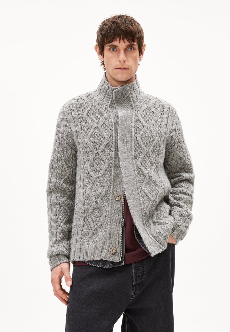 Graue Strickjacke mit hochgeschlossenem Kragen, die über Diamantmuster, eine Knopfleiste und gerippte Bündchen verfügt. Über einem dunklen Oberteil und Jeans getragen.