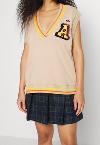 Beige stickad väst med V-ringning och randig orange och gul kant. Har ett broderat bokstaven "A" och är kombinerad med en rutig veckad kjol.