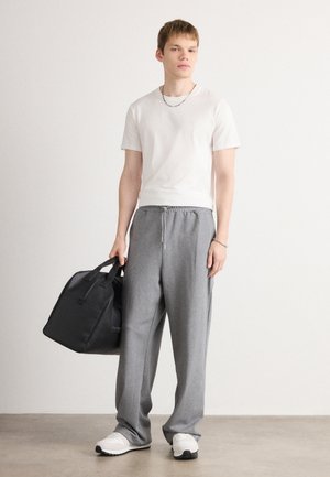 Grijze trainingsbroek, witte aansluitende t-shirt en witte sneakers. Houdt een zwarte totebag vast. Eenvoudig ontwerp, lichtgewicht stof, casual uitstraling.