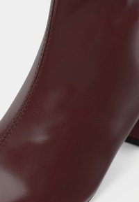Gros plan d'une bottine en cuir marron lisse et brillante avec des coutures visibles sur le côté et un talon bloc sur fond blanc.