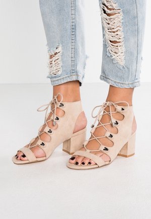 Sandales à talon bloc en daim beige à lacets, portées avec un jean déchiré bleu clair, affichant des orteils vernis en noir.
