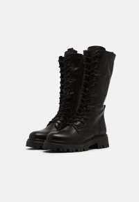 Botas de combate de cuero negro con cordones, de caña alta, suela gruesa y ojales reforzados, que cuentan con una cremallera lateral para mayor comodidad al ponérselas.
