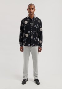 Camisa negra abotonada con patrón floral en blanco y azul, combinada con pantalones gris claro y zapatos negros. Tela con textura y corte relajado.