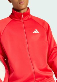 Red zip-up jacket with high collar made of smooth polyester fabric. Features white logo and contrasting shoulder panels.