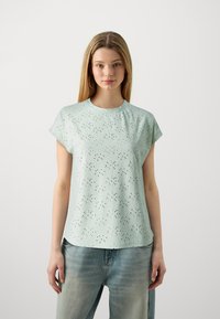 Top corto de manga corta verde claro con un patrón texturizado que presenta cortes, cuello redondo y un ajuste holgado. Combinado con jeans azules desgastados.