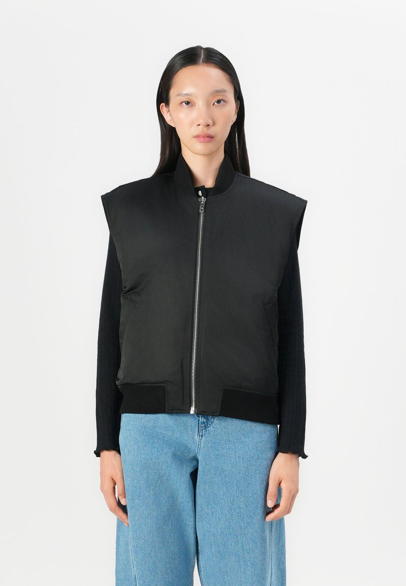 Gilet imbottito nero con chiusura a zip, colletto alto e orlo a costine. Indossato sopra una maglietta nera a maniche lunghe, abbinato a jeans blu chiaro.