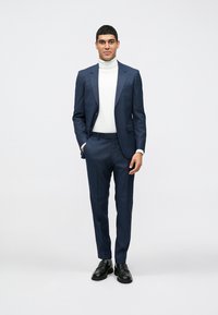 Mornarsko-plavo krojeno odijelo s jednorednim sakoom na jedan gumb i pantalonama ravnog kroja; nošeno preko bijelog rolka; crne elegantne cipele; glatka tekstura.