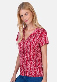 Red short-sleeve blouse featuring a floral pattern in white, v-neckline, button front, and fitted shape paired with blue jeans.