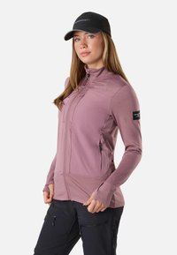 Frau trägt eine mauvefarbene Jacke mit Reißverschluss und Daumenlöchern, schwarze Zip-Hose und eine schwarze Kappe, steht mit den Händen in Hüfthöhe.