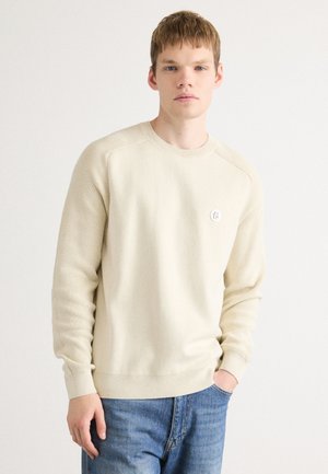 Beige stickad tröja med rund hals, ribbade muddar och fåll, med en liten logotyp patch på vänster bröst, i kombination med blåljeans.