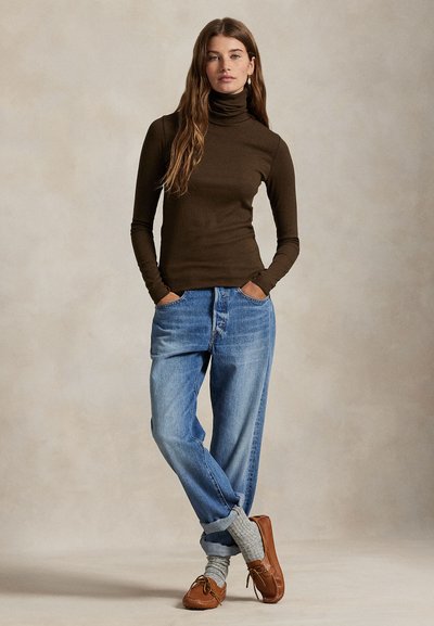 Joven mujer con jersey de cuello alto marrón, vaqueros azules holgados, calcetines grises y mocasines de cuero marrón, de pie con las manos en los bolsillos.