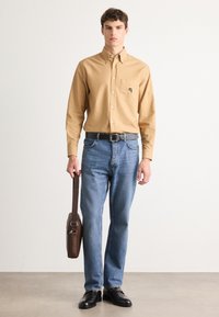 Mann iført beige knappeskjorte, blå jeans, svart belte og sko, holder en brun lærveske i stroppen mot hvit bakgrunn.