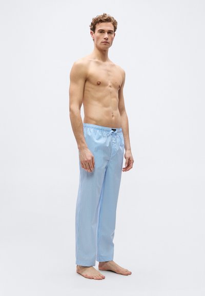 Νέος άνδρας που στέκεται ξυπόλητος, φορώντας σορτς πιτζάμας με φωτεινές γαλάζιες και λευκές ρίγες, χωρίς πουκάμισο, μπροστά σ ε απλό φωτεινό φόντο.