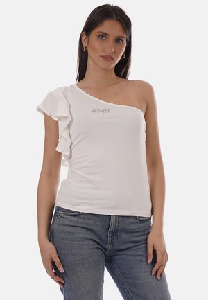 Donna che indossa un top bianco monospalla con dettaglio a balze e orecchini a cerchio dorati, abbinato a jeans di denim blu, in piedi davanti a uno sfondo bianco.