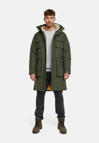 Parka verde imbottito con cappuccio foderato in pelliccia, multiple tasche frontali e una fodera interna gialla, indossato sopra un maglione grigio e jeans scuri.