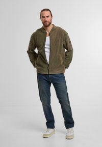 Oliwkowa, rozpinana kurtka z miękkiego materiału, o fasonie bomber i zaokrąglonym kołnierzu, w zestawie z niebieskimi dżinsami i białymi trampkami.