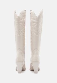 Bottes blanches en cuir montantes avec des têtes angulaires, des bords festonnés et des motifs de couture complexes ; talons arrondis.