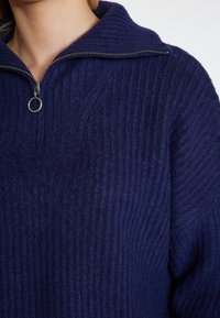 Marineblauwe ribgebreide pullover met een hoge kraag en een halve rits aan de voorkant. Gemaakt van zachte brei-stof, met een gestructureerd oppervlak en een metalen rits.
